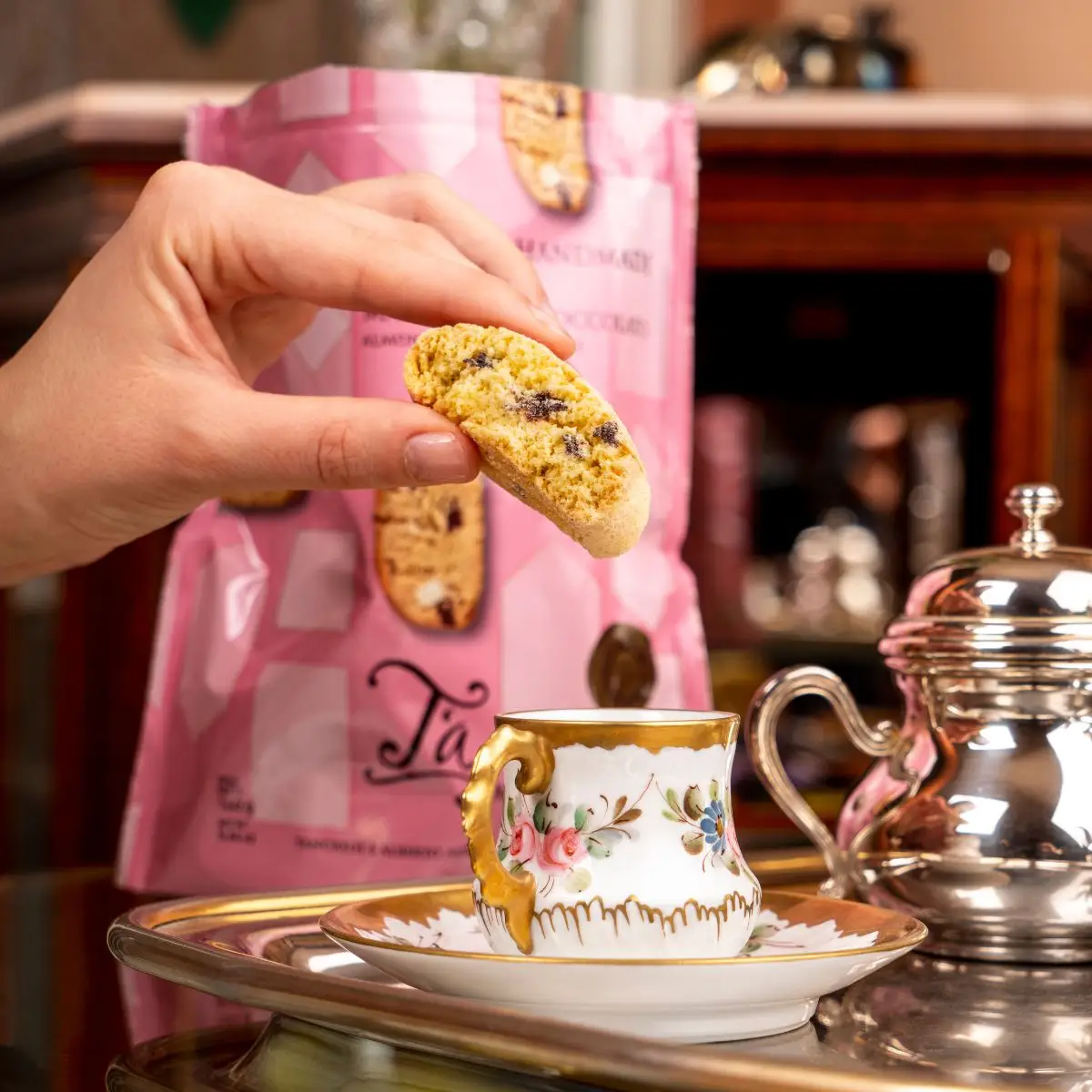 Una mano prende un biscotto con le gocce di cioccolato dal sacchetto rosa T'a, accanto a una tazzina e a un bricco d'argento